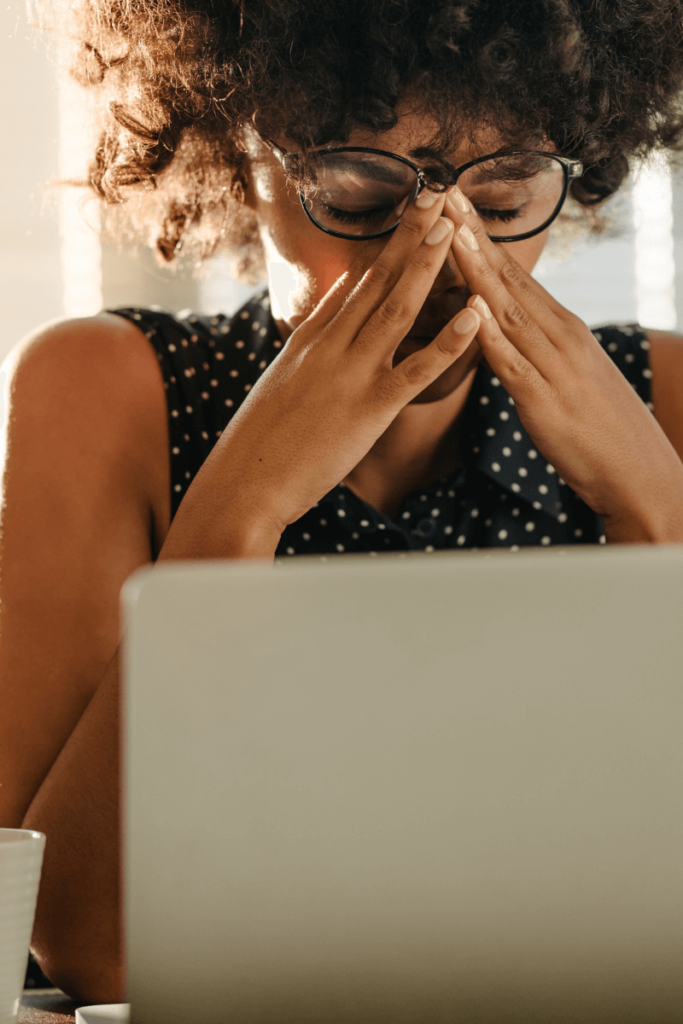 A woman with curly hair and glasses sits at a desk with her hands pressed to her face, appearing stressed. She wears a sleeveless polka dot top and works on a laptop.