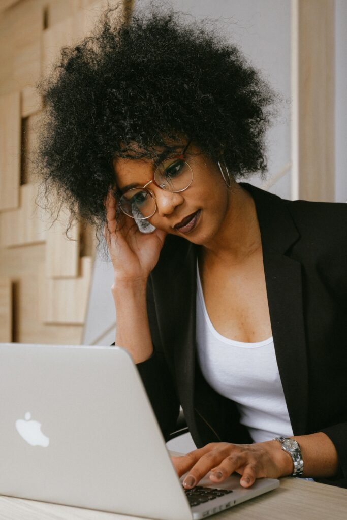 A woman with curly hair and glasses is sitting at a desk, concentrating on her laptop. She rests her head on one hand while typing with the other, dressed in a black blazer and white top.