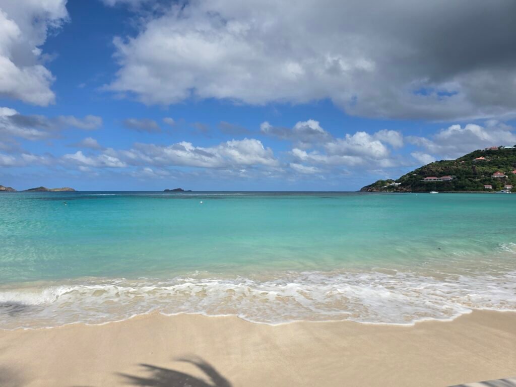 A peaceful, wide-angle view of a tropical beach in Anguilla under a bright blue sky filled with fluffy white and grey clouds. The turquoise Caribbean water gently laps onto a pristine shore of soft, tan sand. In the distance, a lush green hillside dotted with white-roofed villas overlooks the bay, while the faint shadow of a palm frond stretches across the foreground sand.